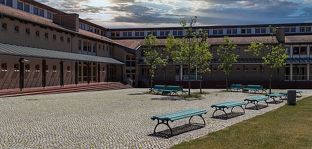 Schulhof mit Schulgebäude und Bänken und Wolken